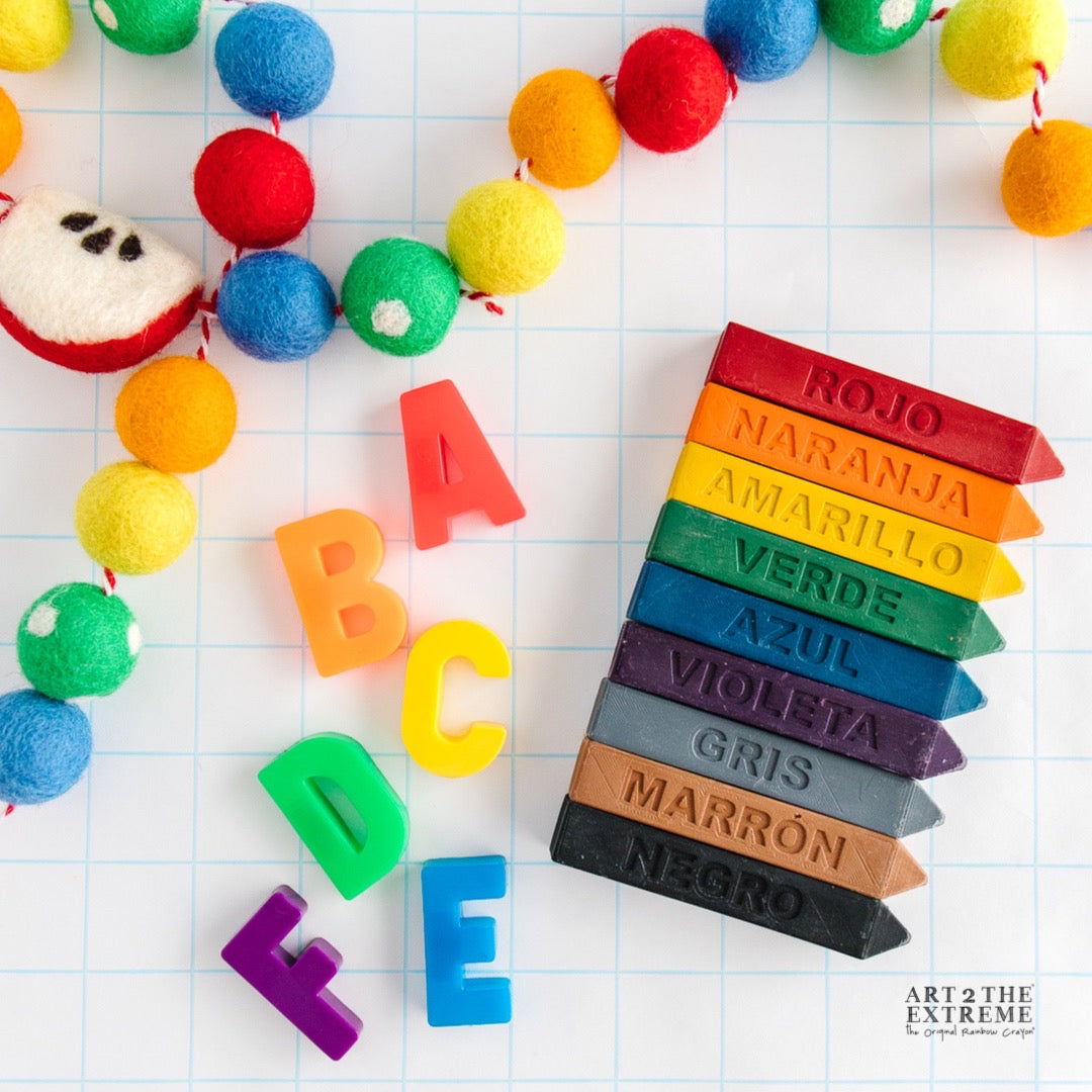 9 spanish colored crayons are on a white background with ABC magnets surrounding them. These crayon gifts for kids are a great way to learn the colors in Spanish and a perfect classroom gift. 