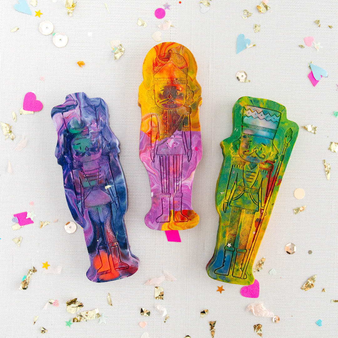 Three Nutcracker-shaped rainbow crayons from Art 2 the Extreme are displayed on a light background, surrounded by scattered confetti and sequins in various shapes, including hearts and stars. Each nutcracker crayon features a unique mix of vibrant colors, such as purple, pink, yellow, green, and blue, with detailed Nutcracker designs. The festive scene adds a whimsical touch, making these crayons a great choice for holiday crafts, Christmas stocking stuffers, and Nutcracker-themed gifts for a girl or boy.
