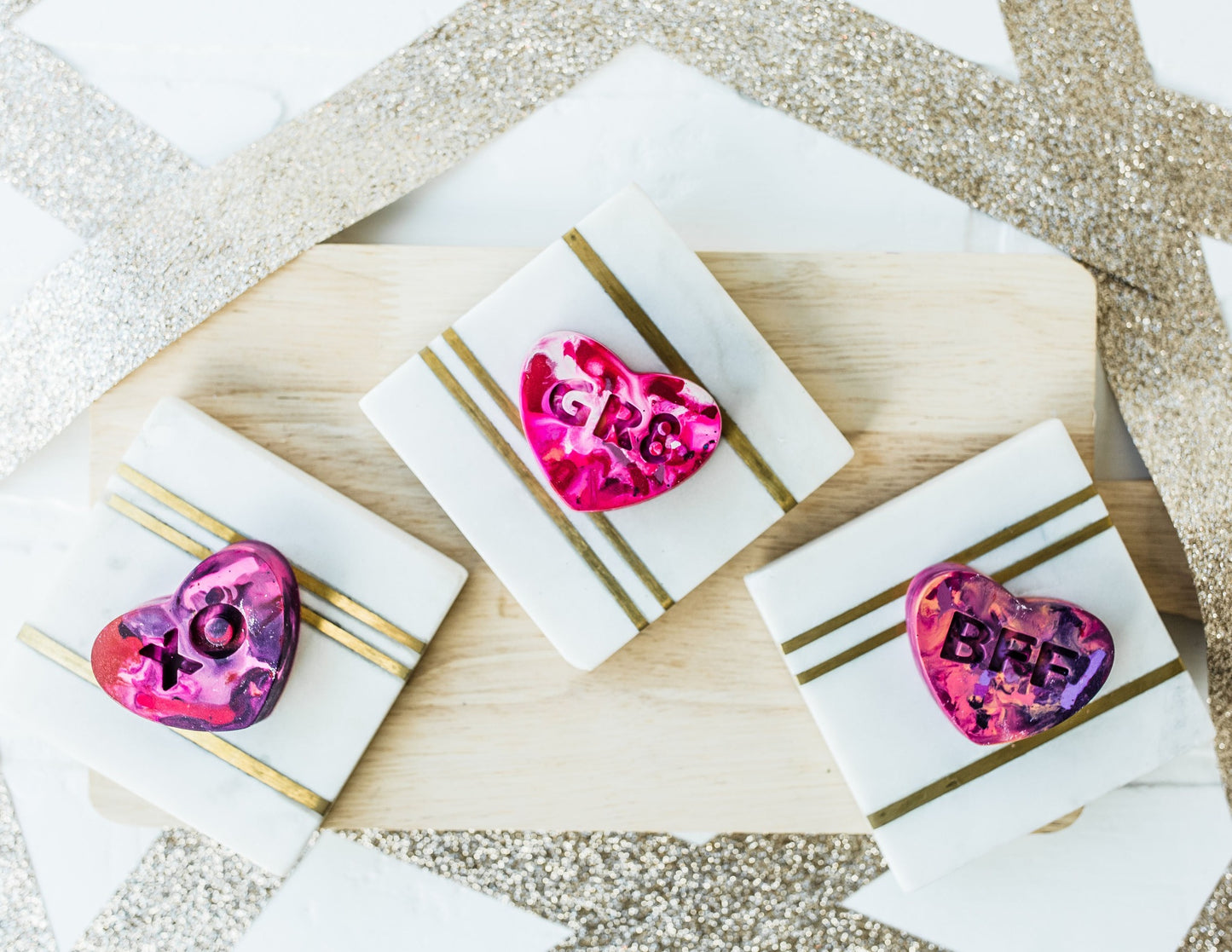 Three multi-colored conversation heart valentine crayons for kids are displayed on a white and gold background with a wooden charcuterie board as a photo prop. Rainbow crayons displayed are a pink/purple/red mixture for Valentine's Day. 