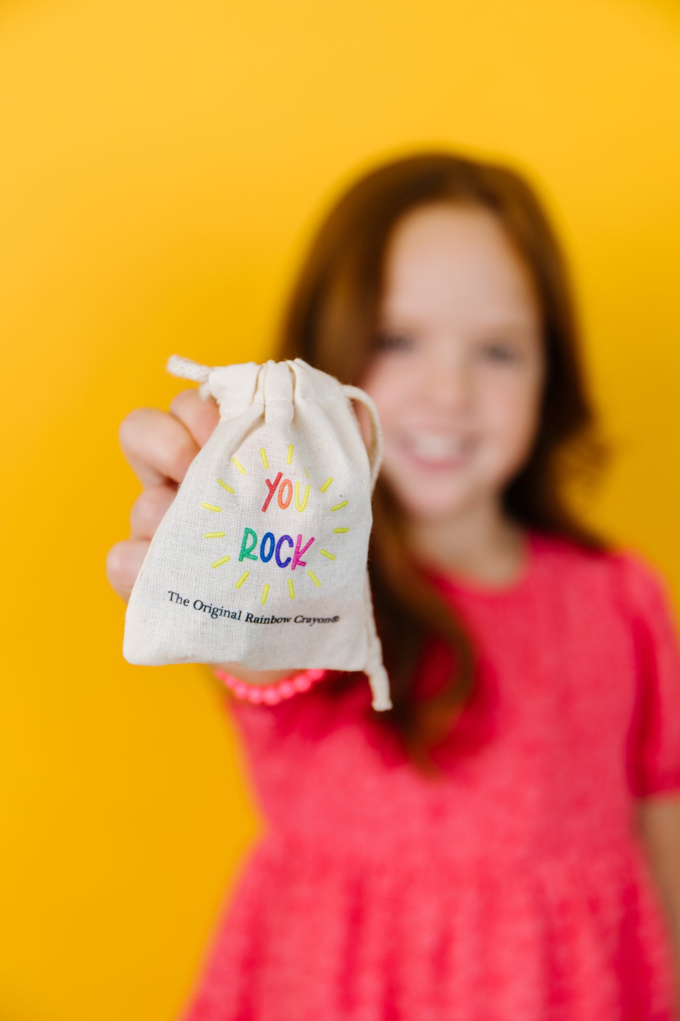 Older child holding crayon rock bag that says YOU ROCK on the outside. THe background is yellow and the bag also says "The Original Rainbow Crayon."