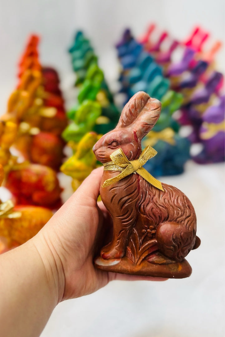 Hand holding pretend chocolate Easter Bunny recycled crayon in brown hues with a gold bow. In the background are 20 rainbow crayon Easter bunny crayons out of focus on a white background. These crayons make cute Easter Basket stuffer gifts for kids.